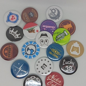 May include: A collection of 15 round, colorful buttons with various designs and text. The buttons feature images and text related to the Fallout video game series, including the Vault-Tec logo, Nuka-Cola, and the Lucky 38 casino.
