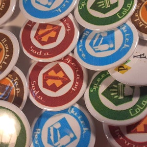 May include: A collection of colorful round buttons with various symbols and text. The buttons feature a variety of designs, including a red button with a cross and the word "Soda", a green button with a tree and the word "Cola", and a blue button with a person and the word "Quick".