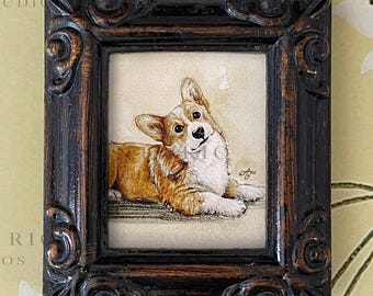 May include: A framed artwork featuring a corgi dog in shades of brown, white, and tan. The dog is lying down with its head tilted. The frame is dark brown with ornate detailing. The background is a light beige.