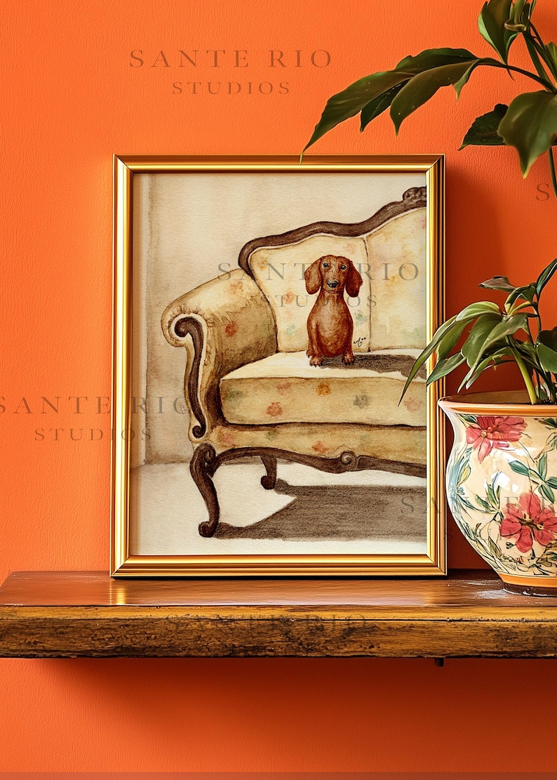 Op de afbeelding: Een ingelijste aquarel van een teckel op een antieke fauteuil met bloemenpatroon. Het kunstwerk zit in een gouden lijst tegen een oranje muur. De tekst "SANTE RIO STUDIOS" is zichtbaar. Thema: dierenkunst.