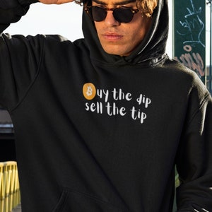 May include: Black hoodie with a front pocket and a hood. The text "Buy the dip sell the tip" is printed on the front, along with a Bitcoin symbol. The person in the image is wearing a black baseball cap and sunglasses.