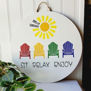 K&ouml;nnte beinhalten: Runder wei&szlig;er Holzschild mit Juteseilaufh&auml;ngung. Es zeigt eine gelbe Sonnengrafik und den Text "Hello Sunshine". Darunter befinden sich vier farbenfrohe Adirondack-Stuhlgrafiken in Rot, Gelb, Gr&uuml;n und Blau mit den Worten "SIT RELAX ENJOY."