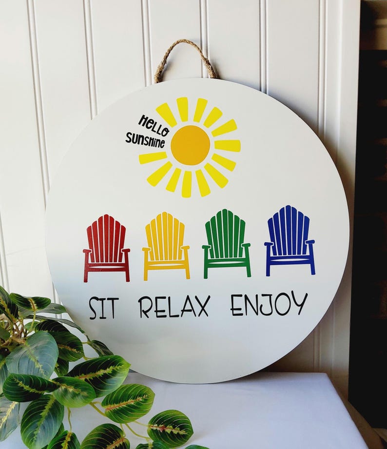 K&ouml;nnte beinhalten: Runder wei&szlig;er Holzschild mit Juteseilaufh&auml;ngung. Es zeigt eine gelbe Sonnengrafik und den Text "Hello Sunshine". Darunter befinden sich vier farbenfrohe Adirondack-Stuhlgrafiken in Rot, Gelb, Gr&uuml;n und Blau mit den Worten "SIT RELAX ENJOY."