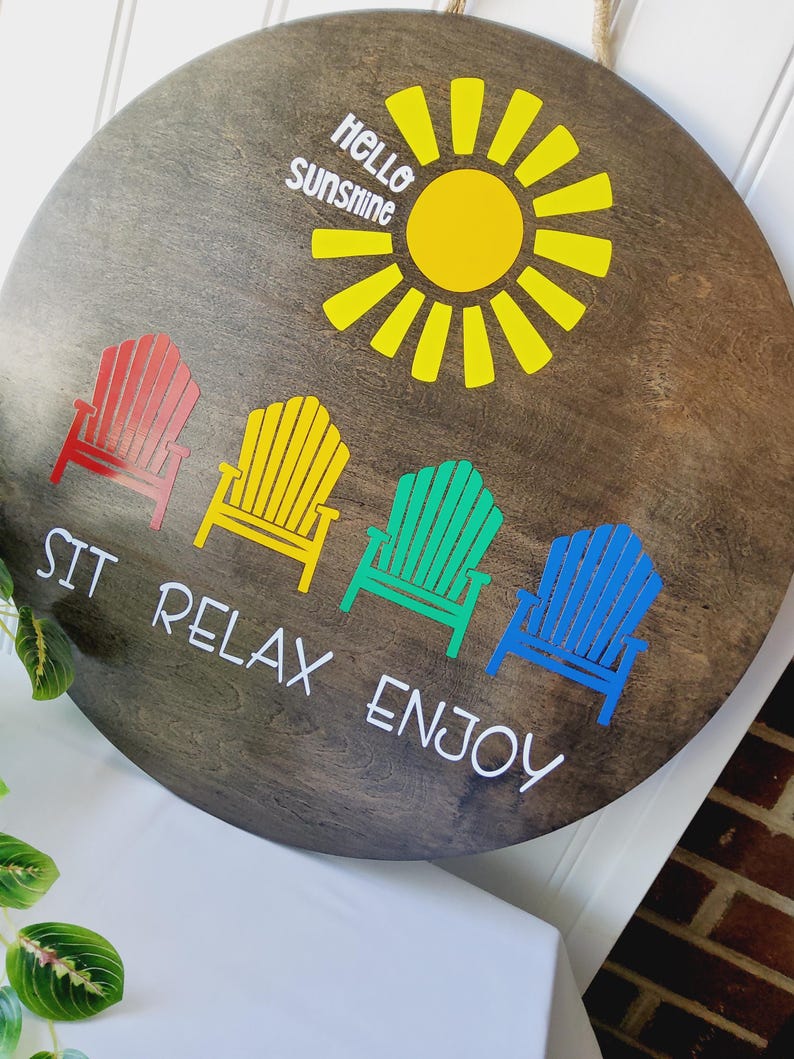 K&ouml;nnte beinhalten: Runder Holzschild mit dunkler Beize, mit einer gelben Sonne und den Worten "Hello Sunshine". Darunter vier farbige Adirondack-St&uuml;hle in Rot, Gelb, Gr&uuml;n und Blau, mit den Worten "Sit Relax Enjoy".