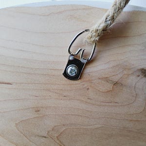 K&ouml;nnte beinhalten: Nahaufnahme eines runden Holzschildes mit nat&uuml;rlicher Holzmaserung. Eine Metallhalterung und eine Schraube sichern ein dickes, gewebtes Seil zum Aufh&auml;ngen. Das Schild hat einen wei&szlig;en Rand.