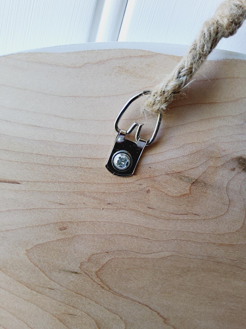 K&ouml;nnte beinhalten: Nahaufnahme eines runden Holzschildes mit nat&uuml;rlicher Holzmaserung. Eine Metallhalterung und eine Schraube sichern ein dickes, gewebtes Seil zum Aufh&auml;ngen. Das Schild hat einen wei&szlig;en Rand.