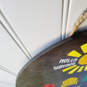 K&ouml;nnte beinhalten: Ein rundes, dunkelbraunes Holzschild mit den Worten "Hello Sunshine" und einer gelben Sonnengrafik. Das Schild zeigt auch die W&ouml;rter "SIT" und "RELAX" mit Stuhlgrafiken. Ein Naturseil ist zum Aufh&auml;ngen befestigt.