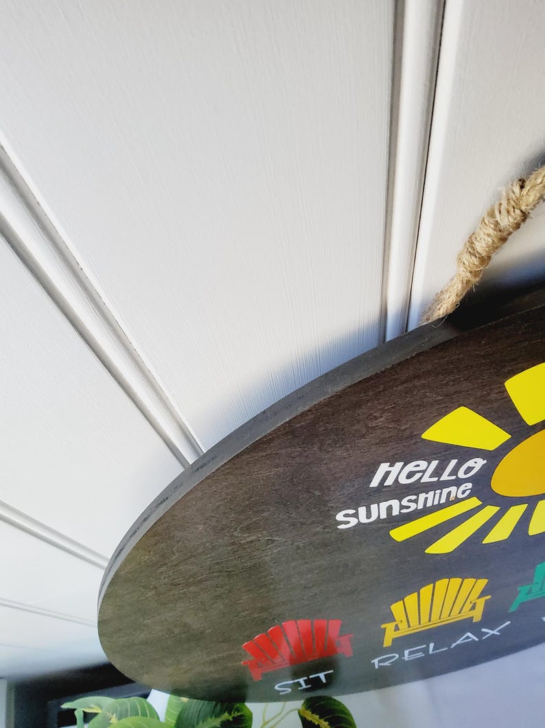 K&ouml;nnte beinhalten: Ein rundes, dunkelbraunes Holzschild mit den Worten "Hello Sunshine" und einer gelben Sonnengrafik. Das Schild zeigt auch die W&ouml;rter "SIT" und "RELAX" mit Stuhlgrafiken. Ein Naturseil ist zum Aufh&auml;ngen befestigt.