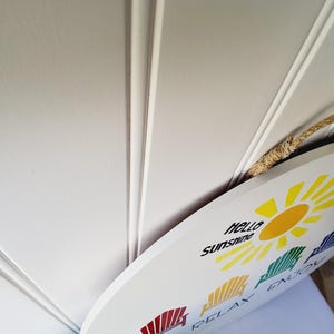 K&ouml;nnte beinhalten: Wei&szlig;es ovales Schild mit den Worten "hello sunshine" und "SIT RELAX ENJOY" in schwarzem Text. Das Schild zeigt eine gelbe Sonnengrafik und farbenfrohe Strandkorbillustrationen. Ein Seilgriff ist angebracht.