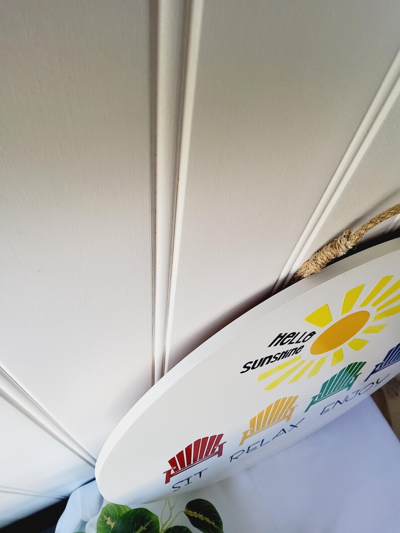 K&ouml;nnte beinhalten: Wei&szlig;es ovales Schild mit den Worten "hello sunshine" und "SIT RELAX ENJOY" in schwarzem Text. Das Schild zeigt eine gelbe Sonnengrafik und farbenfrohe Strandkorbillustrationen. Ein Seilgriff ist angebracht.