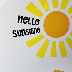 K&ouml;nnte beinhalten: Runder wei&szlig;er Aufsteller mit einem gelben Sonnendesign und den Worten "HELLO SUNSHINE" in Schwarz. Die Sonne hat einen gelben Kreismittelpunkt und gelbe Strahlen. Das Schild hat ein Muscheldesign in der unteren Ecke.