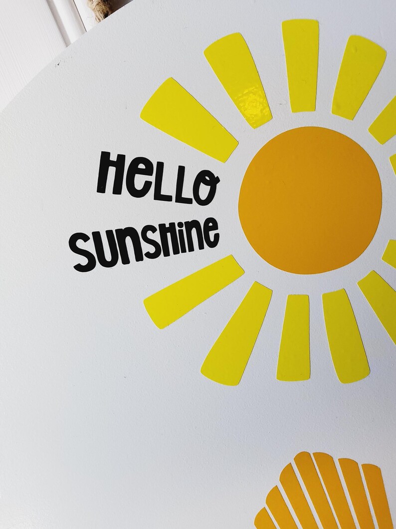 K&ouml;nnte beinhalten: Runder wei&szlig;er Aufsteller mit einem gelben Sonnendesign und den Worten "HELLO SUNSHINE" in Schwarz. Die Sonne hat einen gelben Kreismittelpunkt und gelbe Strahlen. Das Schild hat ein Muscheldesign in der unteren Ecke.