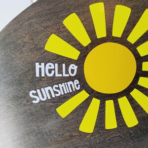 K&ouml;nnte beinhalten: Runder Holzschild mit dunkelbraunem Finish, das ein hellgelbes Sonnendesign zeigt. Die Sonne hat einen gelben Kreismittelpunkt und gelbe Strahlen. Die Worte "HELLO SUNSHINE" sind in Wei&szlig; gehalten und verleihen der Dekoration eine fr&ouml;hliche Note.