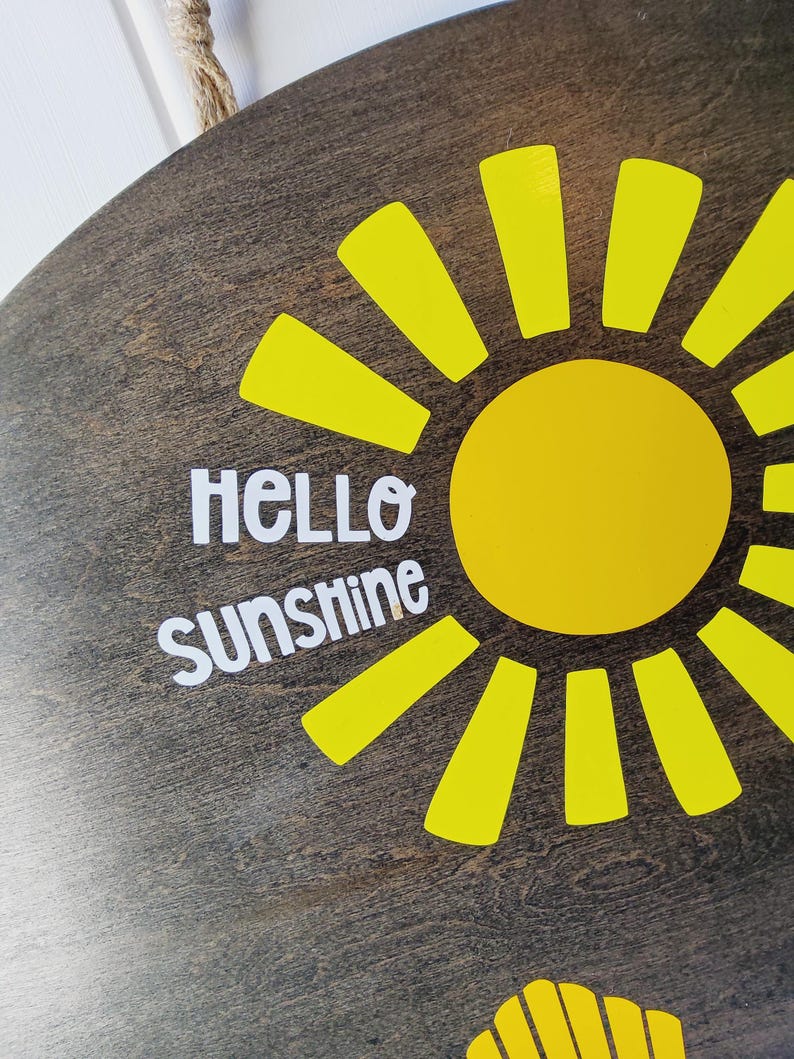K&ouml;nnte beinhalten: Runder Holzschild mit dunkelbraunem Finish, das ein hellgelbes Sonnendesign zeigt. Die Sonne hat einen gelben Kreismittelpunkt und gelbe Strahlen. Die Worte "HELLO SUNSHINE" sind in Wei&szlig; gehalten und verleihen der Dekoration eine fr&ouml;hliche Note.