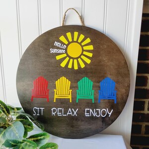 K&ouml;nnte beinhalten: Runder Holzschild mit einer Aufh&auml;ngeschlaufe aus Seil. Es zeigt eine gelbe Sonne und den Text "Hello Sunshine". Darunter befinden sich vier farbige Adirondack-St&uuml;hle in Rot, Gelb, Gr&uuml;n und Blau, mit dem Schriftzug "SIT RELAX ENJOY" darunter.