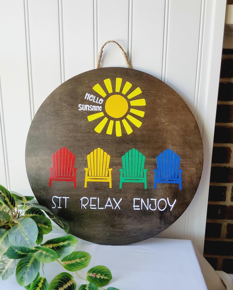 K&ouml;nnte beinhalten: Runder Holzschild mit einer Aufh&auml;ngeschlaufe aus Seil. Es zeigt eine gelbe Sonne und den Text "Hello Sunshine". Darunter befinden sich vier farbige Adirondack-St&uuml;hle in Rot, Gelb, Gr&uuml;n und Blau, mit dem Schriftzug "SIT RELAX ENJOY" darunter.