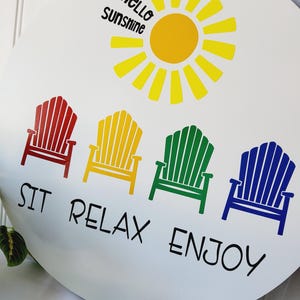 K&ouml;nnte beinhalten: Runder wei&szlig;er Anh&auml;nger mit gelber Sonne und der Aufschrift "Hello Sunshine". Darunter vier farbige Adirondack-St&uuml;hle in Rot, Gelb, Gr&uuml;n und Blau mit den Worten "SIT RELAX ENJOY" in Schwarz.