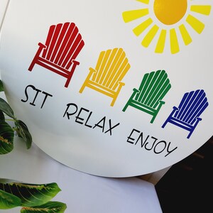 K&ouml;nnte beinhalten: Wei&szlig;es rundes Schild mit einer gelben Sonne und dem Schriftzug "Hello Sunshine". Darunter vier farbige Adirondack-St&uuml;hle in Rot, Gelb, Gr&uuml;n und Blau mit dem Text "SIT RELAX ENJOY".