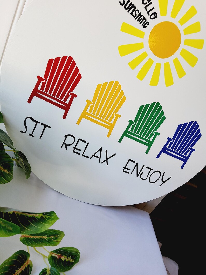 K&ouml;nnte beinhalten: Wei&szlig;es rundes Schild mit einer gelben Sonne und dem Schriftzug "Hello Sunshine". Darunter vier farbige Adirondack-St&uuml;hle in Rot, Gelb, Gr&uuml;n und Blau mit dem Text "SIT RELAX ENJOY".