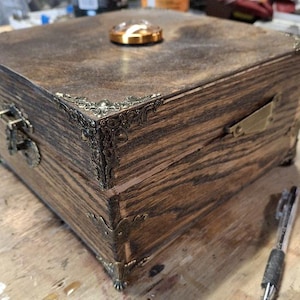 May include: A handcrafted wooden box with a hinged lid, metal corner accents, and a clasp closure. The box has a dark wood grain finish and a small, round, gold-colored object on top. The box is square and appears to be a storage container.
