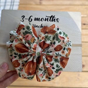 May include: A fabric hair bow with a cream background featuring a pattern of orange pumpkins, pink flowers, and green leaves. The bow is attached to a thin elastic band and is displayed on a light wood sign with the text 3-6 months.