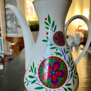 Puede incluir: Cafetera de cerámica blanca con un diseño floral en rojo, verde y azul. El diseño presenta flores y hojas estilizadas.