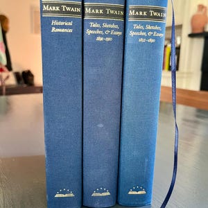Puede incluir: Tres libros de tapa dura de Mark Twain, azul oscuro con letras doradas. Los títulos incluyen "Historical Romances" y "Tales, Sketches, Speeches, & Essays" con fechas de publicación. Una cinta azul como marcapáginas es visible.