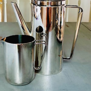 May include: A polished silver-tone metal coffee pot with a tall cylindrical body, a long spout, a lid with a small knob, and a large handle, positioned next to a smaller matching silver-tone metal creamer pitcher with a handle and a pouring spout.