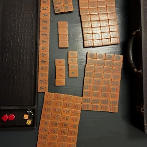 Könnte beinhalten: Eine Sammlung von Vintage-Mahjong-Steinen, die auf einer dunklen Oberfläche angeordnet sind. Die Steine sind hauptsächlich beige mit schwarzen und roten Markierungen und weisen verschiedene Symbole und Zeichen auf. Rote und grüne Würfel sind sichtbar.
