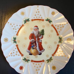 May include: White ceramic plate with a scalloped edge. The center of the plate features a festive illustration of Santa Claus holding a Christmas tree and a bag of gifts. The illustration is surrounded by a gold and red border with holly and snowflakes.