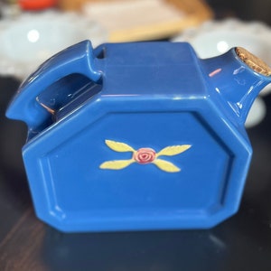 May include: A blue ceramic teapot with a cork stopper and a yellow and pink floral design. The teapot has an octagonal shape and a handle.