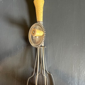 May include: A vintage hand-crank egg beater with a yellow speckled handle and three metal beaters. The beaters are attached to a metal gear that is turned by the handle.