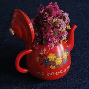 May include: A red ceramic teapot with a floral design, filled with dried purple and yellow flowers. The teapot has a curved handle and a spout. The lid is open, revealing the flowers inside. The teapot is decorated with hand-painted yellow and blue flowers.