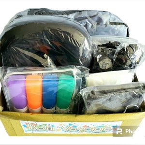 May include: A cardboard box with the words "Bon Voyage" printed on the front. The box is filled with various travel items, including a black neck pillow, a black eye mask, a white cloth, and four colorful highlighters.