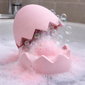 May include: A pink, egg-shaped bath bomb holder with a cracked top, filled with pink foam and surrounded by bubbles. The holder is in a bathtub filled with soapy water, creating a playful and bubbly scene.