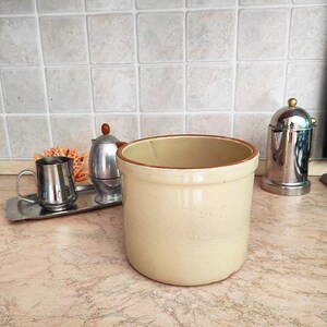 Può includere: Un barattolo di ceramica color crema con un bordo marrone si trova su un piano di lavoro. Il barattolo è vuoto e pronto per essere riempito.