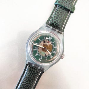 Puede incluir: Un reloj Swatch con caja transparente y esfera verde con números romanos. El reloj tiene una correa de cuero verde texturizado con costuras blancas. La esfera revela el mecanismo interno.
