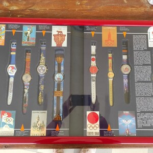 Puede incluir: Una vitrina enmarcada de relojes Swatch que conmemora los Juegos Olímpicos. Los relojes presentan diseños y logotipos coloridos de varios Juegos Olímpicos, incluyendo Tokio 1964, Seúl 1988 y Barcelona 1992. La vitrina también incluye carteles e información sobre la historia de los Juegos Olímpicos.