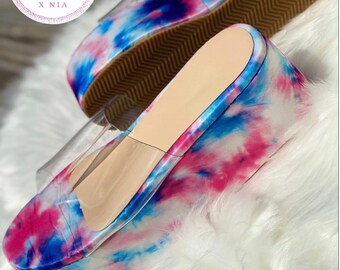 May include: A pair of clear platform sandals with a tie-dye pattern in shades of pink, blue, and white. The sandals have a thick platform sole and a clear strap across the top.