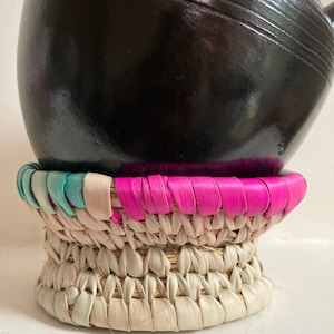 Puede incluir: Una tetera de cerámica negra con una base de paja tejida. La base está decorada con ribetes rosa, blanco y azul.