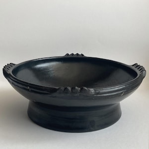 May include: A black, handcrafted ceramic bowl with a wide, shallow basin and a raised pedestal base. The bowl features decorative, ridged edges and small, pointed accents around the rim. The dark color and simple design suggest a rustic or artisanal aesthetic.