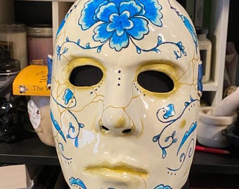 May include: A decorative mask with a cream-colored base, adorned with blue floral designs and gold accents. The mask features eye and mouth openings, with a smooth, glossy finish. The design includes a large flower on the forehead and smaller floral patterns.