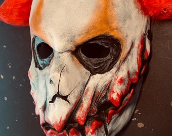 May include: A white and red clown mask with black eye sockets and a cracked face. The mask has orange accents and red hair on top. The mask is designed to look scary and is likely for Halloween or costume use.