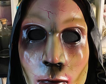 May include: A full-face Halloween mask with a pale complexion, dark eye sockets, and a black hood. The mask has red crack-like markings and a red-tinted mouth. The overall design gives a spooky and unsettling appearance, perfect for costume parties or haunted houses.