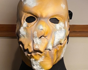 May include: A full-face mask featuring a weathered, mustard-yellow finish with distressed white patches and deep cracks across the surface. The mask has two dark, oval-shaped eye openings and a jagged, textured appearance with a black strap on the side.
