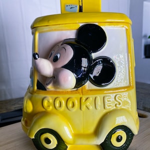 May include: Yellow ceramic cookie jar shaped like a school bus with Mickey Mouse driving. The bus has the word "COOKIES" written on the side.