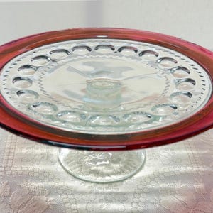 May include: A clear glass cake stand with a ruby red rim. The stand features a circular design with a series of small, decorative indentations around the edge. The base is a clear, rounded pedestal. The cake stand is designed to display baked goods.