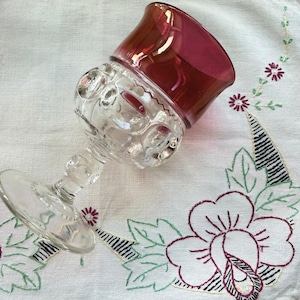 May include: A clear glass goblet with a ruby red bowl. The goblet features a decorative, patterned stem and bowl. The glass is resting on a white tablecloth with embroidered floral designs in shades of purple, green, and black.