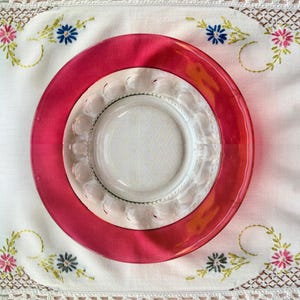 May include: A stack of three plates on a white embroidered tablecloth. The top plate is a vibrant red, the middle plate is clear with a decorative edge, and the bottom plate is white. Floral embroidery decorates the tablecloth.