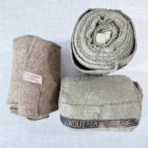 May include: Three rolls of khaki-colored fabric, possibly bandages or wraps. One is tightly rolled, another is partially unrolled, and the third is folded. Labels with text are visible on two of the rolls. The fabric appears to be a coarse weave.